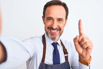 Middle age shopkeeper man wearing apron make selfie over isolated white background surprised with an idea or question pointing finger with happy face, number one
