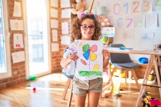 Beautiful toddler wearing glasses and unicorn diadem standing holding draw smiling at kindergarten