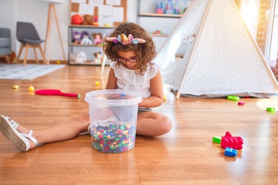 Beautiful toddler wearing glasses and unicorn diadem sitting on the floor playing with building blocks at kindergarten