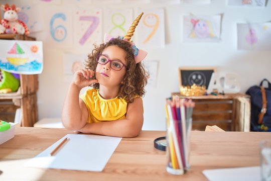 Beautiful toddler wearing glasses and unicorn diadem sitting on desk at kindergarten with hand on chin thinking about question, pensive expression. Smiling with thoughtful face. Doubt concept.