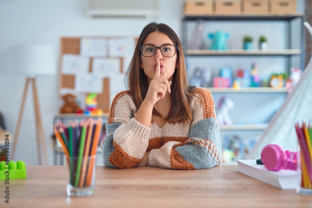 Poster Young beautiful teacher woman wearing sweater and glasses sitting on desk at kindergarten asking to be quiet with finger on lips. Silence and secret concept.