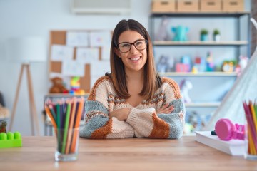 Young beautiful teacher woman wearing sweater and glasses sitting on desk at kindergarten happy...