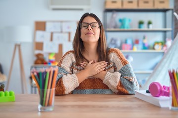 Young beautiful teacher woman wearing sweater and glasses sitting on desk at kindergarten smiling...