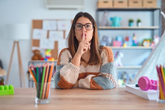 Young Beautiful Teacher Woman Wearing Sweater And Glasses Sitting On Desk At Kindergarten Asking To Be Quiet With Finger On Lips. Silence And Secret Concept.