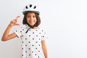 Beautiful child girl wearing security bike helmet standing over isolated white background smiling and confident gesturing with hand doing small size sign with fingers looking and the camera. Measure 