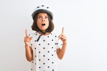 Beautiful child girl wearing security bike helmet standing over isolated white background amazed and surprised looking up and pointing with fingers and raised arms.