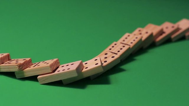 Wooden dominoes are lined up for a domino show, and quickly knocked down. They stand against a green background with pronounced shadows.