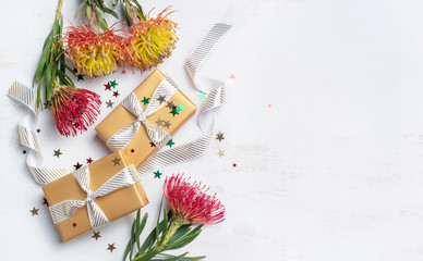 Australian native red and yellow Waratah/Proteas on a rustic white wooden background scattered with glitter and gold gifts that are wrapped in luxury white ribbon.