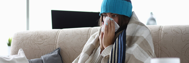 Portrait of of girl sitting in living room covered in blanket and suffering from common cold. Woman...