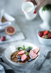 Healthy breakfast with granola, yogurt, fruits, berries on a white plate in white plate. Selective focus. A woman pours milk into granola.