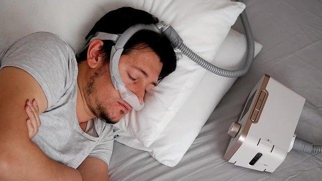 Close-up of healthy young adult man wearing under the nose nasal mask ( CPAP mask ) and using CPAP machine for sleeping smooth without snoring.