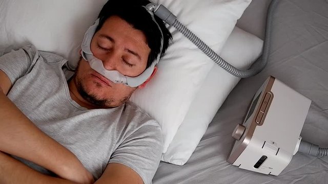 Close-up of healthy young adult man wearing under the nose nasal mask ( CPAP mask ) and using CPAP machine for sleeping smooth without snoring.
