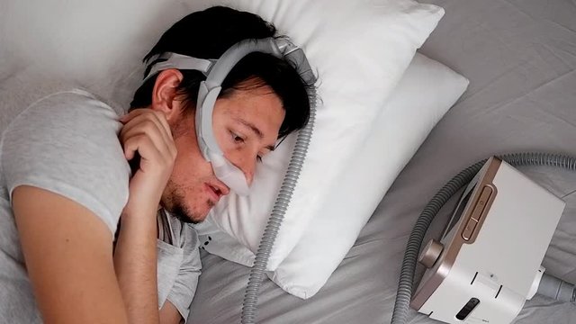 Close-up of healthy young adult man wearing under the nose nasal mask ( CPAP mask ) and using CPAP machine for sleeping smooth without snoring.