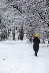 woman in black winter coat and yellow hat walking by snowed city park