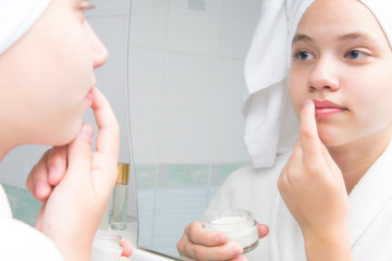 a girl with a white towel on her head and in a bathrobe, in the bathroom, near the mirror, puts cream on her lips to soften