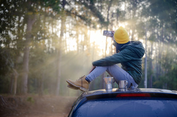 Woman traveller enjoy coffee time with taking shot setie on her owns roof of the car with scenery view of the forest and light of mist morning in background