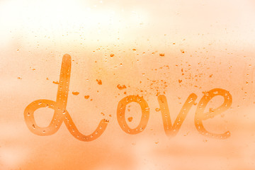 The romantic inscription love on the orange or pink evening or morning window glass with drops 