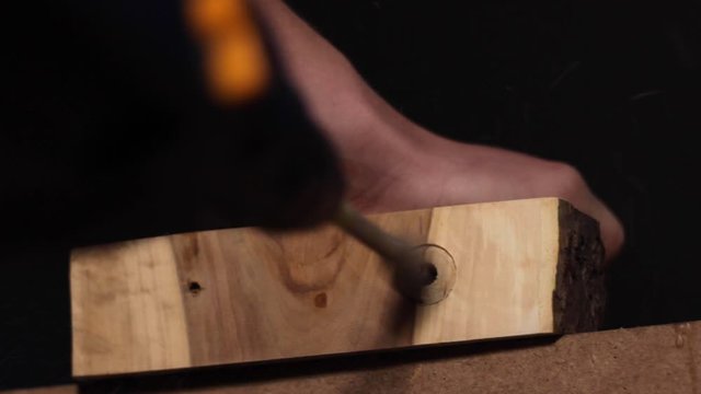 Close up of a carpenter using a special drill to create a large hole in a thick slab of processed, smooth wood, holding it steady with his hand
