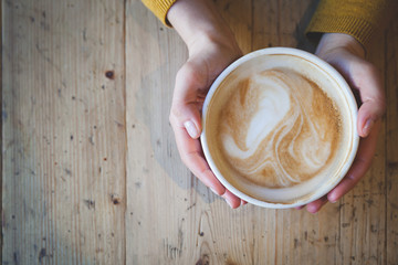 A young girl sits at a wooden table and holds a large cup of latte coffee. Directly above....