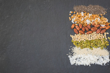 Top view on rows with organic granola with nuts, coconut flakes, sesame seeds, pumpkin seeds, cacao beans and chia seeds. A mixture of different types of beans and grains on a black stone background