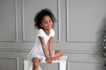 A portrait of an African ballerina in a white gymnastic costume sits on a wooden chair. © evelinphoto