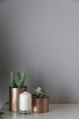 Close up  copper vase  with artificial plant inside and white candle in glass on cream spray-painted working table with  gray painted wall in the background /apartment interior /copy space