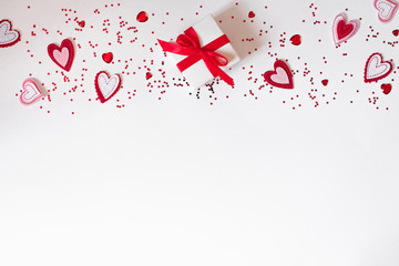 Flat lay composition of a gift box with a red ribbon and felt hearts and confetti on a white background