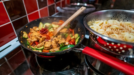  Close up and selective focus of a spicy chicken dish cooking in a non-stick wok on the oven hob and freshly cooked pasta draining in a stainless steel colander