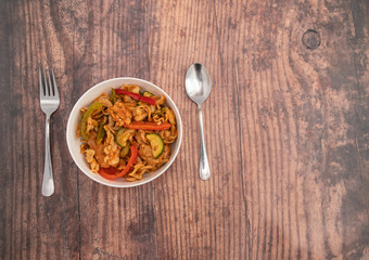  Flat lay of a white bowl of a spicy chicken pasta dinner with a stainless-steel fork and spoon on a wooden background with copy space