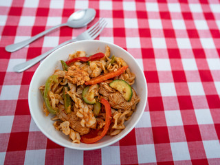  Selective focus on the contents of a spicy chicken and pasta dish with a stainless-steel fork and spoon on a red and white checked table cloth