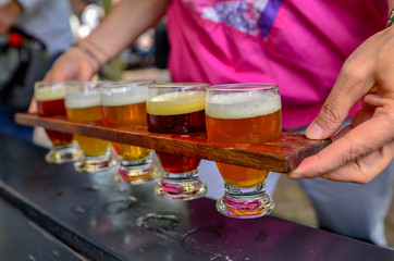 Craft beer tasting: five glasses with beers of different colors and flavors. A wooden cup holder holds the glasses in a row. Two Caucasian woman's hands hold him up.