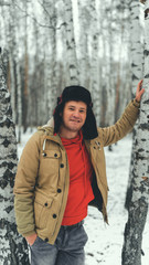 Fototapeta na wymiar Man standing in forest in winter day. Serious young male wearing beige jacket over red hoodie and earflap hat standing on snowy forest and looking away