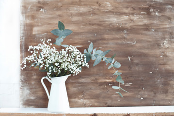jug with white flowers and eucalyptus branches on the background of a shabby wooden wall.