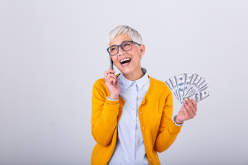 Excited beautiful senior woman talking on her smartphone and holding money, cashback. Finances and people concept, senior woman holding hundreds of dollar money banknotes