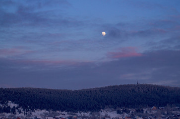The moon in the sky with clouds over the evening, winter forest and the village. The sky with clouds, a round moon illuminates the coniferous forest and houses of the city. Beautiful sky and moonlight