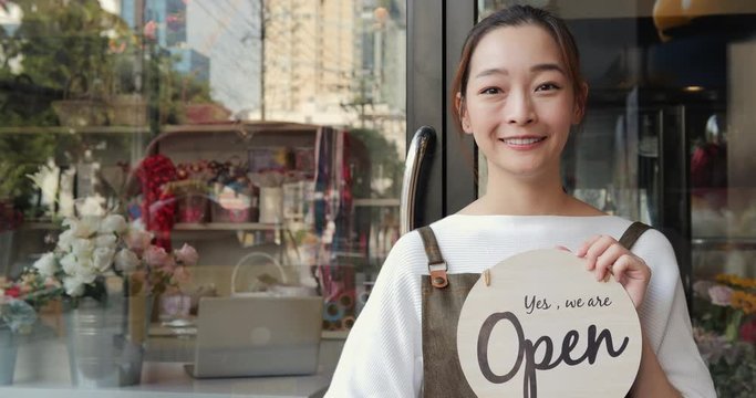 Startup woman holding open sign in hand and looking to camera with smile. She carrying open sign at storefront.