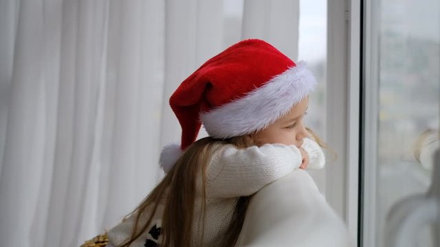 A little girl in a Santa hat sits by the window and waits for Christmas and New Year.