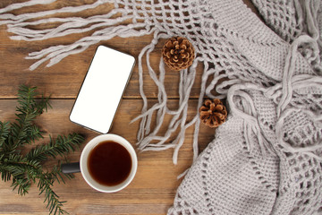 Fototapeta na wymiar flat lay background with a mug of tea, white smart phone with blank screen, shawl with fringe, coniferous branches and dried orange cinnamon on wood, concept winter mood, winter is love, copy space