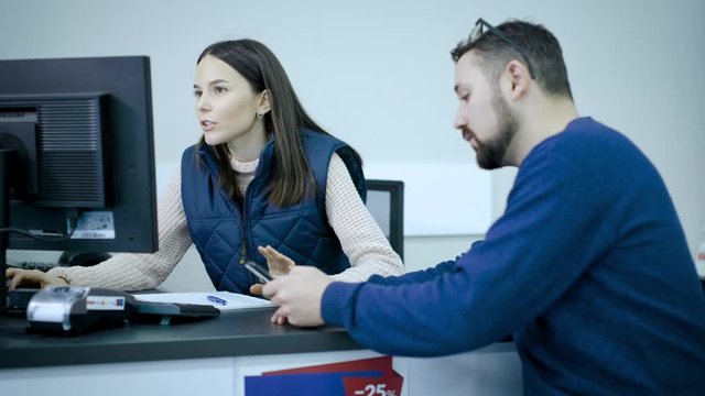 Girl, a company manager in a uniform sitting at a computer communicates with a client. Client, a young guy uses his smartphone to purchase goods. Series.