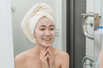 Asian girl in a mask with a towel in front of the mirror in the bathroom. the concept of self-care, moisturizing and cleansing the skin. care of youth and beauty