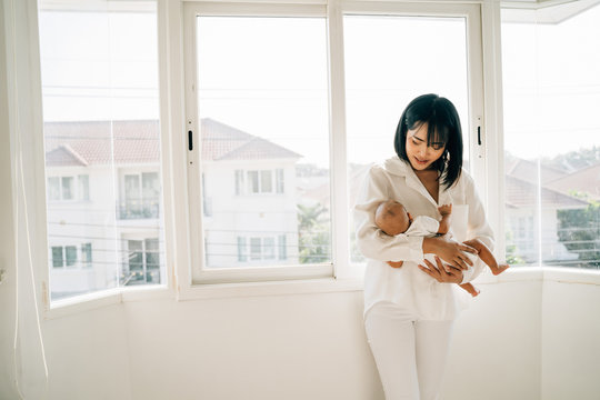 Happy caring young Asian mum in white wear holding own infant standing near window at home