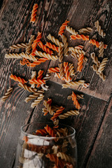 multi-colored spiral raw pasta crumbled out of a glass jar for reusable storage of products on a dark wooden tabletop