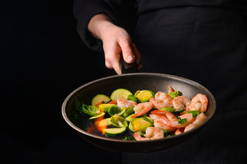 Chef cooks with fire in a pan shrimp with vegetables. On a black background, restaurant service