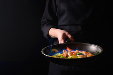 Professional chef cook on a dark background prepares seafood, shrimp with fire and vegetables. Roasting and cooking veggie food. Black background, Space for design.