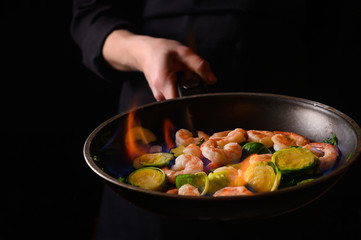 Chef cooks with fire in a pan shrimp with vegetables. On a black background, restaurant service