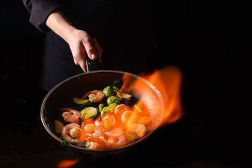 Professional cook cooks with fire. Cooking Cooking seafood, healthy vegetarian food on an open fire on a dark background. Hotel service photo background. Horizontal view.