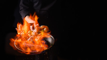 Seafood and their preparation, a professional chef cooks shrimps with vegetables with fire. On a black background, a banner, cooking and recipes. Restaurant and hotel service concept.