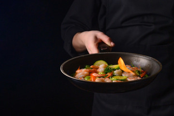 Professional chef cook on a dark background prepares seafood, shrimp with fire and vegetables. Roasting and cooking veggie food. Black background, Space for design.