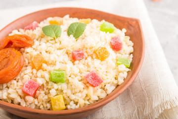 Bulgur porridge with dried apricots and candied fruits in clay bowl on a gray concrete background and linen textile. Side view, selective focus.