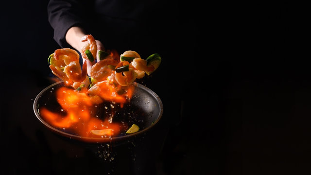 Seafood and their preparation, a professional chef cooks shrimps with vegetables with fire. On a black background, a banner, cooking and recipes. Restaurant and hotel service concept.